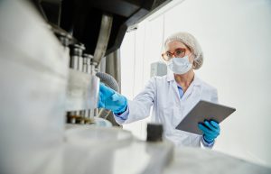 Shot from pill press machine of female process technician in sterile classroom clothing holding digital tablet while inspecting manufacturing equipment at pharmaceutical factory, copy space